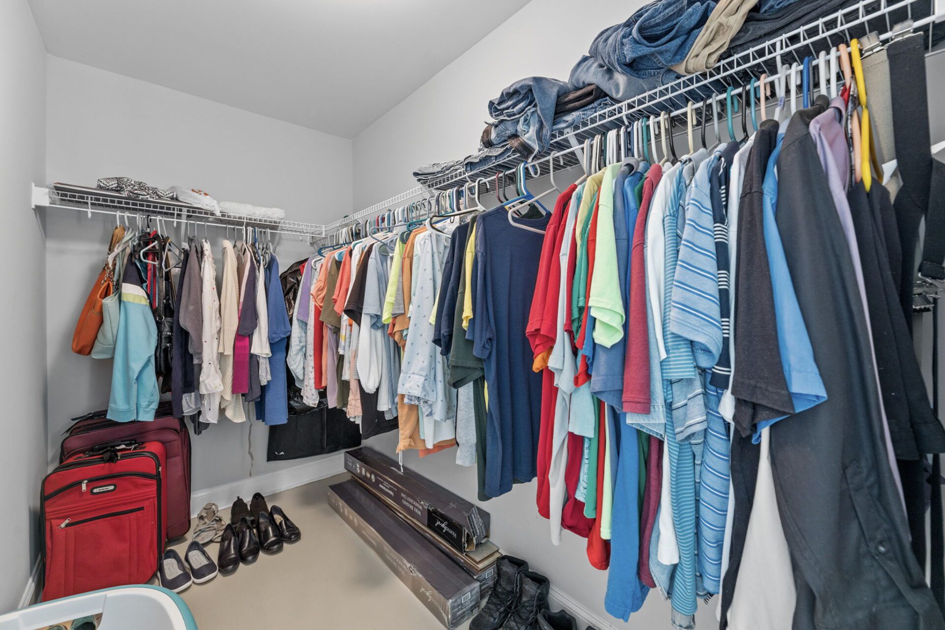 A neatly organized walk-in closet with clothing items hung on racks and shoes placed on the floor. luggage and storage bins are stored neatly beneath hanging clothes.